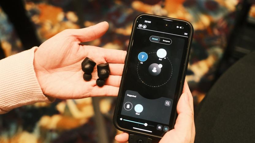 A pair of black earbuds on a person's palm, with their other hand holding up a phone with the OrCam Hear app on its screen.