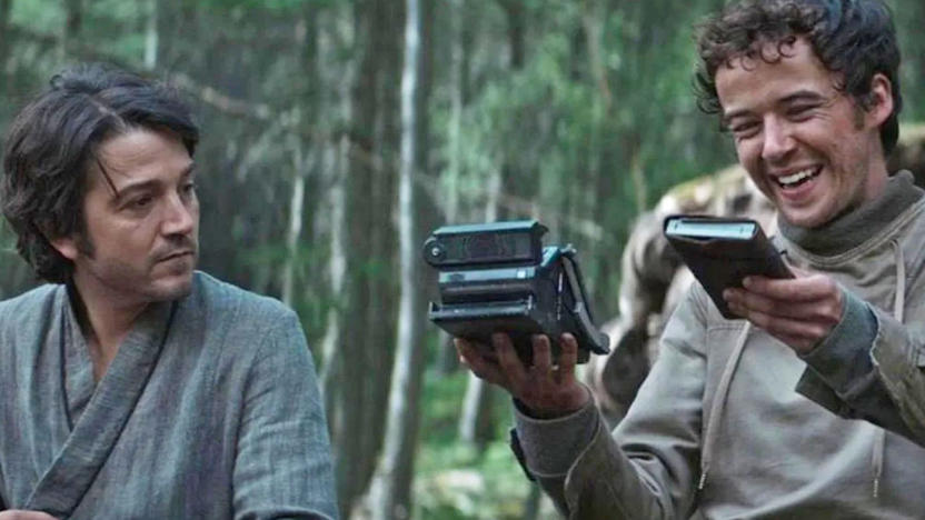 A still image from the tv series 'Andor' showing the main character sitting next to someone who's holding a device that appears to look like a folding polaroid camera. 