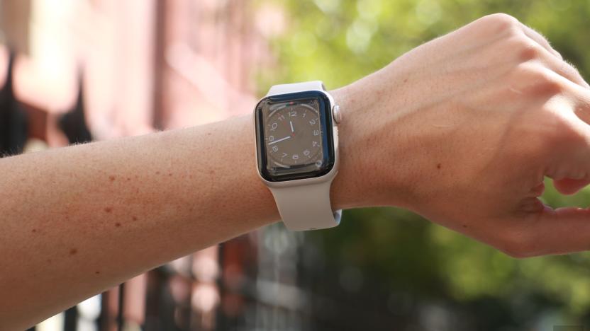 The Apple Watch SE (2022) on a person's wrist held up in front of a red building and a tree.