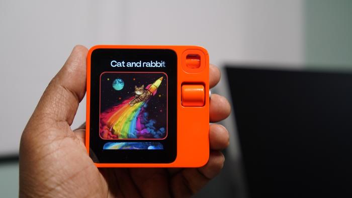 A hand holding a small square orange device with a display.